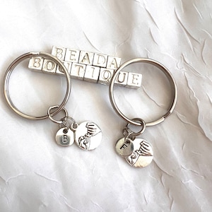 May include: Two silver keychains with interlocking hands charms and letter charms. The keychains are labeled "B" and "F". The keychains are on a white background with the text "BEADA BOUTIQUE" in silver.