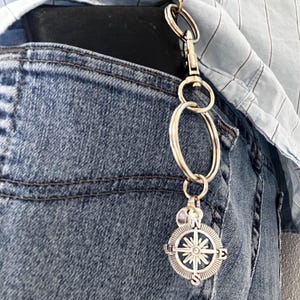 May include: A silver-toned metal keychain featuring a circular compass charm with cardinal directions N, S, E, and W engraved on the rim. The charm hangs from a series of interconnected metal rings and a lobster clasp attached to a black wallet.