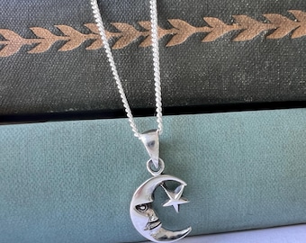 May include: A silver necklace with a crescent moon pendant featuring a face and a star. The necklace is on a stack of books with a green cover.