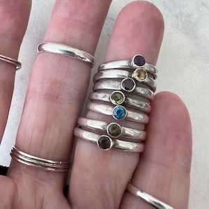 May include: A stack of silver rings with colorful gemstones. The rings are stacked on a finger and feature a variety of colors, including purple, yellow, green, blue, and brown.