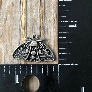 May include: Silver charm in the shape of a moth with moon phases and stars. The charm is approximately 2.5 cm wide.