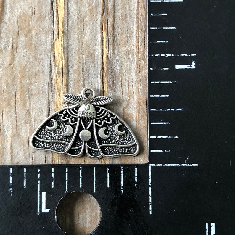 May include: Silver charm in the shape of a moth with moon phases and stars. The charm is approximately 2.5 cm wide.