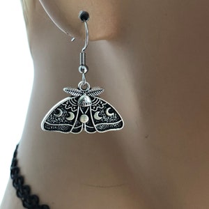 May include: Silver dangle earrings featuring a moth design with crescent moons and a circle in the centre.