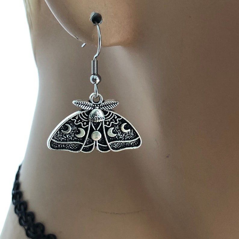 May include: Silver dangle earrings featuring a moth design with crescent moons and a circle in the centre.