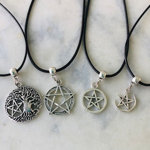 May include: Four silver-toned pendant necklaces with black cords. The pendants feature a tree of life with a pentagram, a pentagram, a pentagram within a circle, and a crescent moon with a pentagram. The necklaces are displayed on a white marble surface.