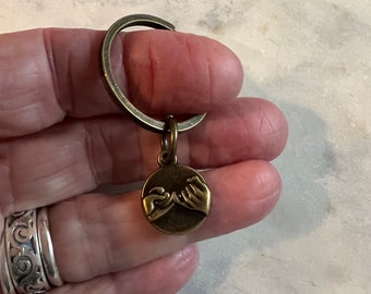 May include: A bronze keychain with a round charm featuring two hands making a pinky promise. The keychain is attached to a silver key ring.