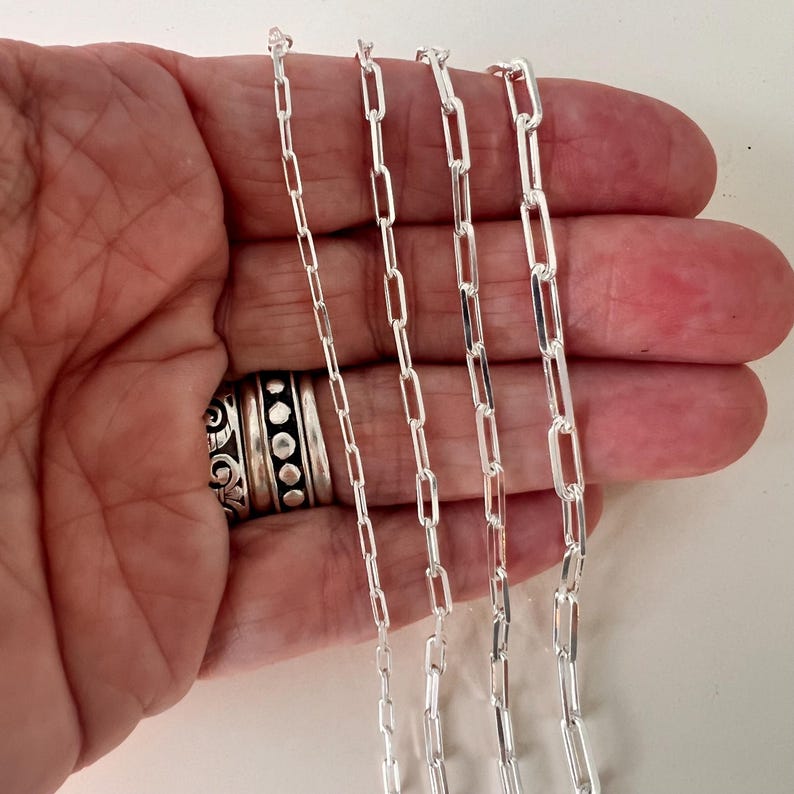 May include: Four silver chain necklaces of varying lengths, displayed on a hand. The chains are made of small, rectangular links.