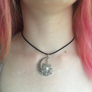 May include: A silver-colored pendant necklace featuring a crescent moon and sun design. The moon has an ornate, openwork pattern, and the sun hangs from the moon's center. The necklace has a black cord.