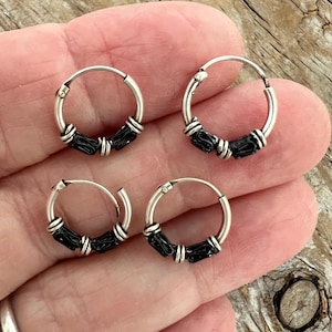 May include: Four small silver hoop earrings with black accents. Each earring has a circular design with a silver hoop and a series of black beads. The earrings are displayed on a light-colored surface.