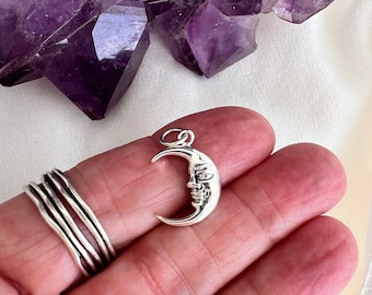 May include: A silver crescent moon charm with a face on it. The charm is being held in a hand.
