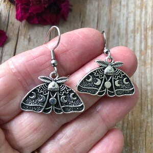May include: Silver dangle earrings featuring a moth design with crescent moons and stars. The moth is detailed with a textured finish.