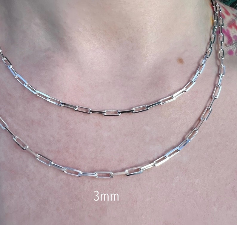 May include: Two silver chain necklaces, one with a 3mm width, layered on a person's neck. The chains are made of small, rectangular links.