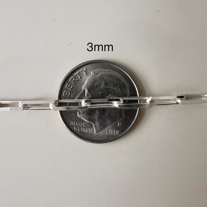 May include: Close-up of a silver chain with a 3mm width, measured against a US dime coin. The chain is made of linked rectangular pieces.