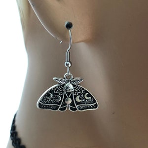 May include: Silver dangle earrings featuring a moth with crescent moons and a sun on its wings.