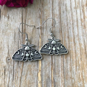 May include: A pair of silver earrings featuring a moth design with crescent moons and a full moon. The earrings are hanging from silver hooks.
