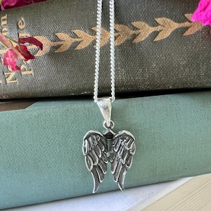 May include: A silver necklace with a pendant featuring a pair of detailed angel wings. The wings are silver and have a textured finish.