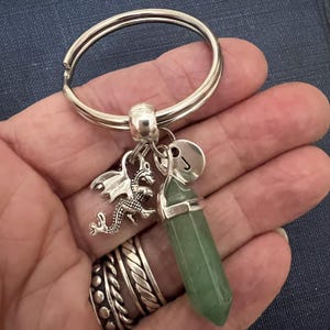 May include: Silver keychain with a green crystal pendant, a dragon charm, and a circular charm with the letter "J". The keychain is attached to a silver split ring.