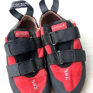 May include: A pair of Boreal climbing shoes in red and black with two black hook-and-loop straps across the top. The shoes feature a black rubber toe cap, a yellow sole, and a black pull tab on the heel with the white text BOREAL printed on it.