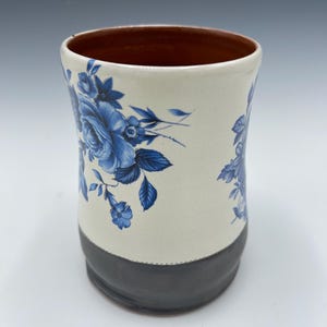 May include: A ceramic mug with a cream-colored body, adorned with blue floral patterns. The base is dark gray, and the interior is brown. The mug has a cylindrical shape and is suitable for beverages.