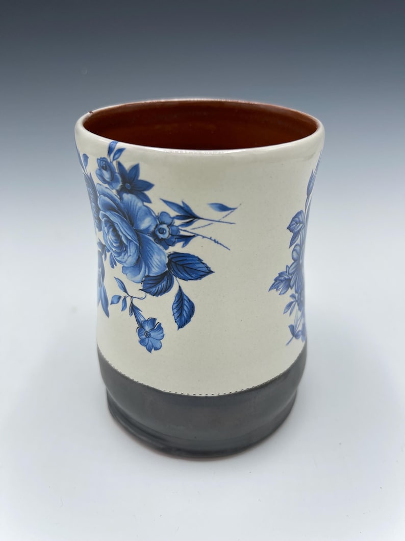 May include: A ceramic mug with a cream-colored body, adorned with blue floral patterns. The base is dark gray, and the interior is brown. The mug has a cylindrical shape and is suitable for beverages.