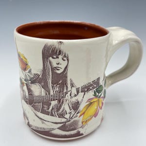 May include: Cream-colored ceramic mug featuring a sepia-toned image of a woman playing a guitar. The mug has a brown interior and a curved handle. Yellow and pink floral accents are also present on the mug.