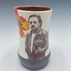 May include: A white ceramic mug with a black handle and the text "But First, Coffee" in a bold, black font. The mug is set against a blurred background, suggesting a cozy setting.