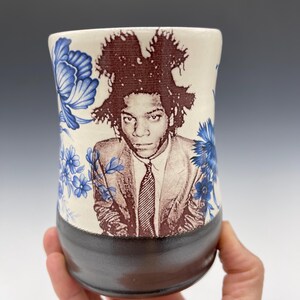 May include: A ceramic tumbler featuring a portrait of a person in a suit and tie, with a dark brown base and a cream-colored upper section. The upper section is decorated with blue floral patterns.