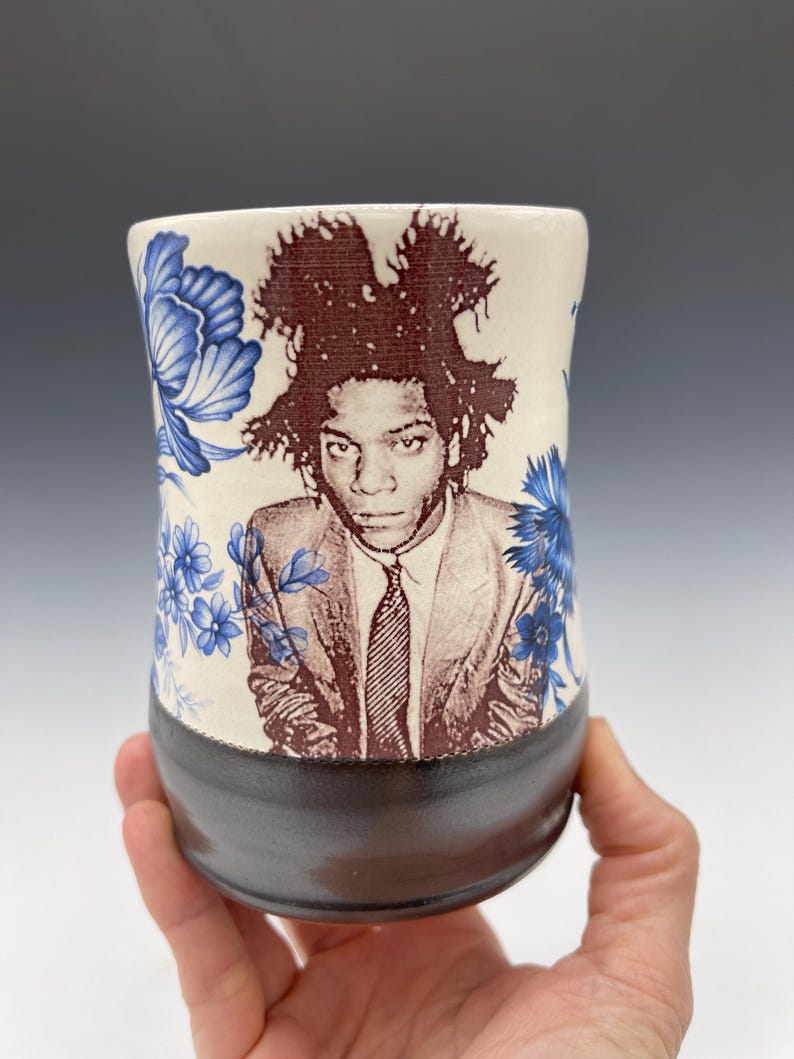 May include: A ceramic tumbler featuring a portrait of a person in a suit and tie, with a dark brown base and a cream-colored upper section. The upper section is decorated with blue floral patterns.
