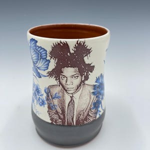 May include: A ceramic mug featuring a portrait of a person in a suit with a tie. The mug has a white background with blue floral patterns. The bottom of the mug is gray, and the inside is brown.