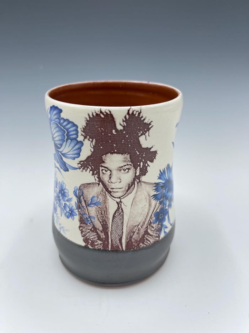 May include: A ceramic mug featuring a portrait of a person in a suit with a tie. The mug has a white background with blue floral patterns. The bottom of the mug is gray, and the inside is brown.