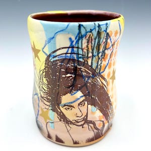 May include: A ceramic tumbler featuring a dark brown screen-printed portrait of a person with long hair. The surface is decorated with light blue and yellow abstract shapes, orange polka dots, and gold stars against a cream-colored background.