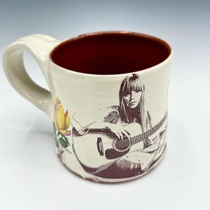 May include: A ceramic mug with a cream-colored exterior and a deep red interior. The mug features a dark brown graphic print of a person playing an acoustic guitar, accompanied by a yellow rose illustration on the side and a large handle.