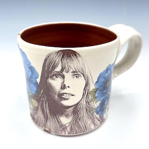 May include: A ceramic mug with a cream-colored exterior and a dark reddish-brown interior. The front features a detailed monochromatic portrait of a person with long hair, flanked by vibrant blue floral illustrations on both sides.