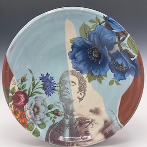 May include: A ceramic plate with a light blue background, featuring a floral design with blue flowers and a portrait of a person. The rim is a reddish-brown color. The plate has a unique artistic design.