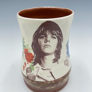 May include: A ceramic mug with a brown rim and base, featuring a black and white portrait of a person. The mug is decorated with floral patterns in red, blue, and green. The mug is approximately 4 inches tall.