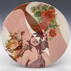 May include: A decorative ceramic plate featuring a woman's portrait and floral arrangements. The plate has a pink and white background with brown accents. The floral arrangements include red and yellow roses.
