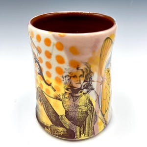 May include: A ceramic tumbler with a deep maroon interior and a cream-colored exterior. The surface features a graphic print of a person with wavy hair, orange polka dots, and a large yellow butterfly wing extending along the side of the vessel.