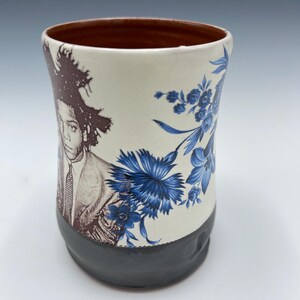 May include: A ceramic mug with a brown interior, a cream-colored exterior, and a dark gray base. The mug features a portrait and blue floral design. The portrait is of a person with dark hair and a suit. The floral design is of blue flowers.