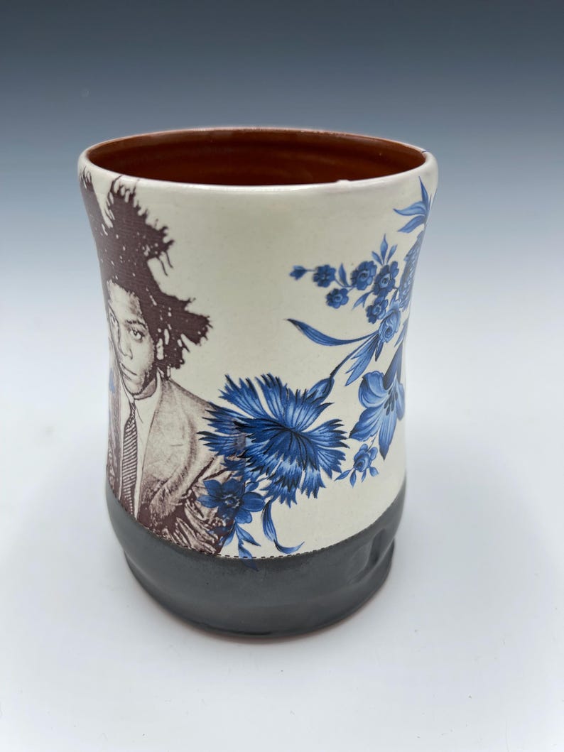 May include: A ceramic mug with a brown interior, a cream-colored exterior, and a dark gray base. The mug features a portrait and blue floral design. The portrait is of a person with dark hair and a suit. The floral design is of blue flowers.