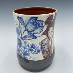May include: A ceramic mug with a brown interior and a dark gray base. The mug features a blue floral pattern and a sepia-toned portrait. The mug is cylindrical and has a slightly flared rim.