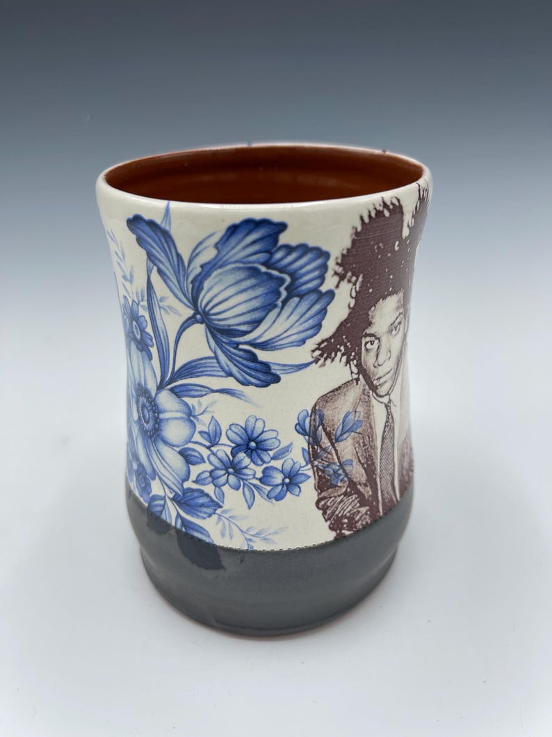 May include: A ceramic mug with a brown interior and a dark gray base. The mug features a blue floral pattern and a sepia-toned portrait. The mug is cylindrical and has a slightly flared rim.