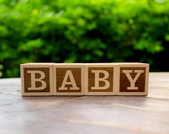 Puede incluir: Cuatro bloques de madera que deletrean la palabra "BABY". Los bloques son de color marrón claro con las letras en marrón más oscuro. Los bloques están sobre una superficie de madera con un fondo verde borroso.