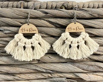 Puede incluir: Un par de pendientes de macramé de color crema con círculos de madera que dicen "He is Risen" en letras marrones.