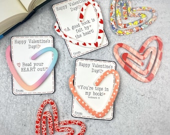 May include: Four heart-shaped bookmarks with different designs and messages. The bookmarks are made of paper and have a white background with red, pink, and blue accents. The messages on the bookmarks include "Happy Valentine's Day!" "A good book is felt by the heart!" "Read your HEART out!" and "You're tops in my book!"