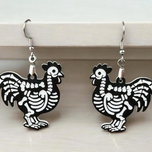 Puede incluir: Un par de pendientes de pollo esqueleto de plata. Los esqueletos de pollo son negros y blancos y tienen un dise&ntilde;o detallado.