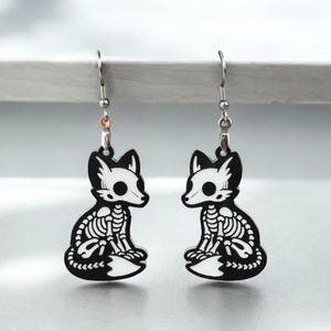 Puede incluir: Un par de pendientes de plata con un dise&ntilde;o de zorro esqueleto en blanco y negro. El zorro est&aacute; sentado con la cola enroscada alrededor de su cuerpo.
