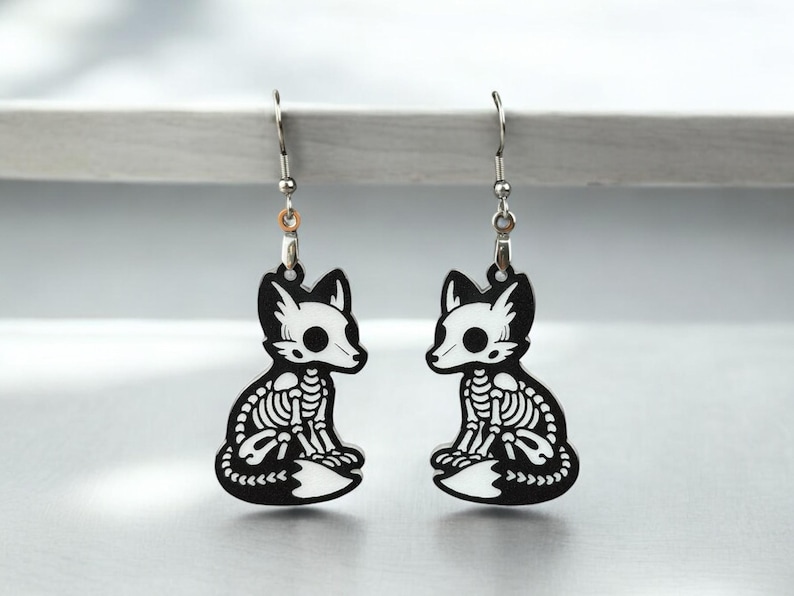 Puede incluir: Un par de pendientes de plata con un dise&ntilde;o de zorro esqueleto en blanco y negro. El zorro est&aacute; sentado con la cola enroscada alrededor de su cuerpo.