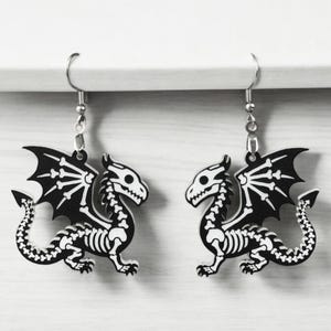 Puede incluir: Un par de pendientes de drag&oacute;n esqueleto en blanco y negro. Los pendientes est&aacute;n hechos de metal y tienen un cierre de gancho plateado.