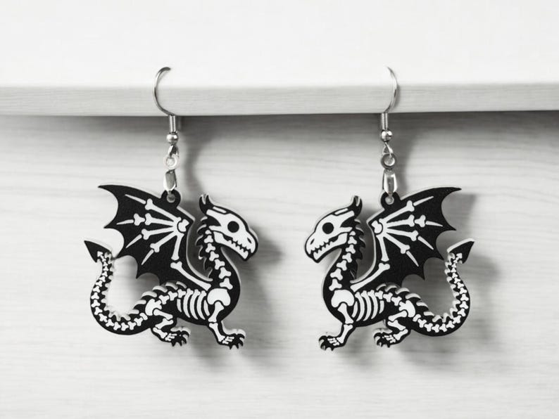 Puede incluir: Un par de pendientes de drag&oacute;n esqueleto en blanco y negro. Los pendientes est&aacute;n hechos de metal y tienen un cierre de gancho plateado.