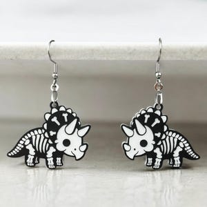 Puede incluir: Un par de pendientes de plata con un dise&ntilde;o de triceratops esqueleto en blanco y negro. Los pendientes est&aacute;n hechos de metal y tienen un dise&ntilde;o colgante.